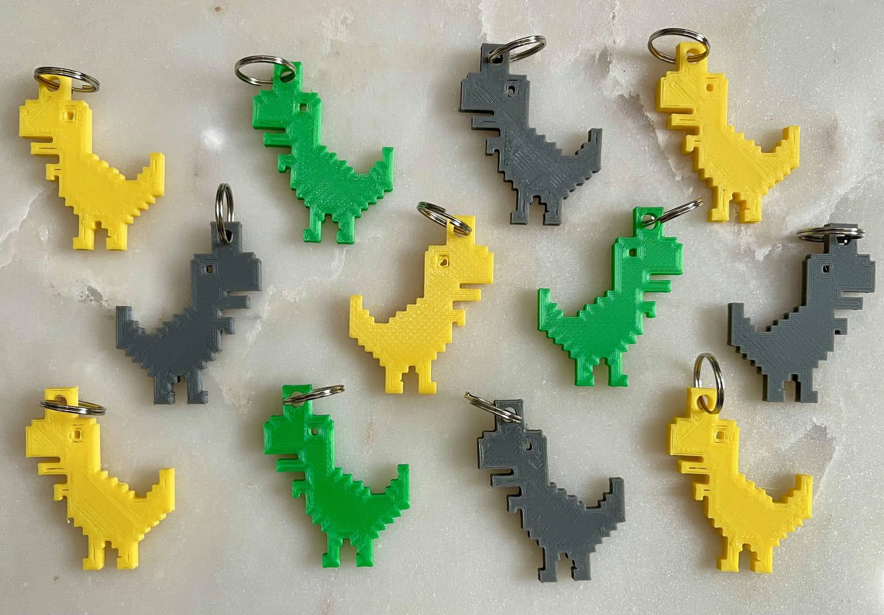 Photo of 12 8-Bit Dino Keyrings in varying colours (Grey, Yellow and Green) on a white marble background.